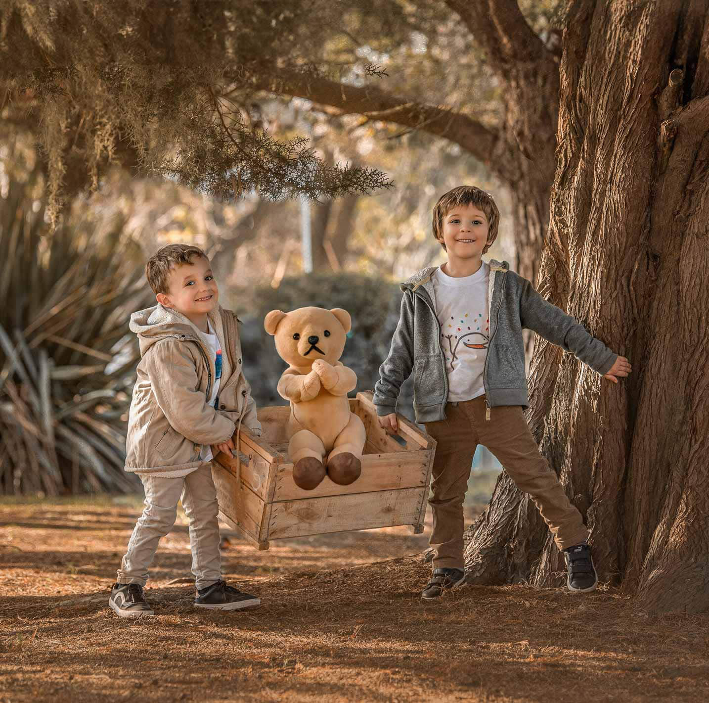 Dos niños sonrientes jugando con un oso de peluche en un cajón de madera, bajo un árbol en un entorno natural – fotografía infantil por Caro Roig.